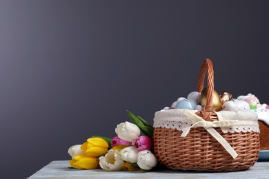 Photo of Wicker basket with festively decorated Easter eggs and beautiful tulips on white wooden table against dark grey background. Space for text
