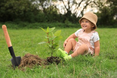 Photo of Cute happy baby girl near young green tree and trowel in garden