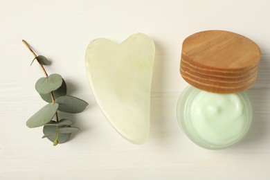 Photo of Jade gua sha tool, eucalyptus branch and jar of cream on white wooden table, flat lay