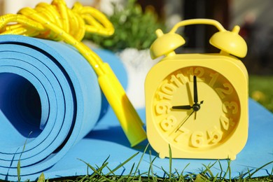 Alarm clock, fitness mat and skipping rope on green grass outdoors, closeup. Morning exercise