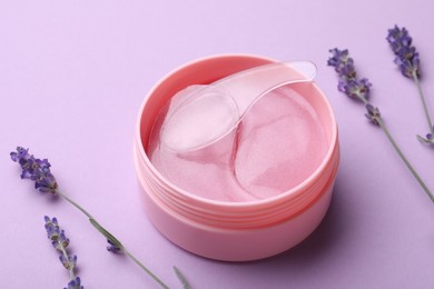 Photo of Under eye patches in jar with spatula and lavender flowers on lilac background. Cosmetic product