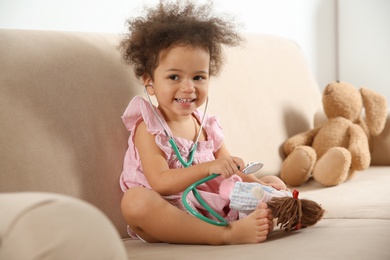 Cute African American child imagining herself as doctor while playing with stethoscope and doll on couch at home