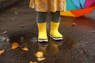 Little girl wearing yellow rubber boots standing in puddle outdoors, closeup