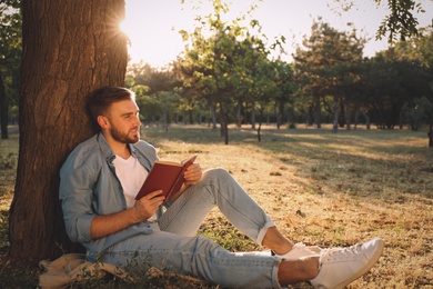 Young man reading book on green grass near tree in park