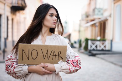 Sad woman in embroidered dress holding poster No War on city street. Space for text