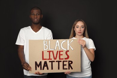 Young woman and African American man holding sign with phrase Black Lives Matter on dark background. Racism concept