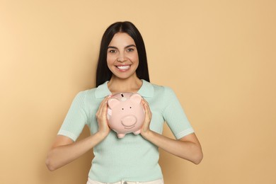 Happy young woman with ceramic piggy bank on beige background