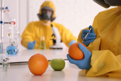 Scientist in chemical protective suit injecting orange at laboratory, closeup
