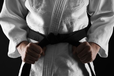 Man in keikogi tying black belt on dark background, closeup. Martial arts uniform