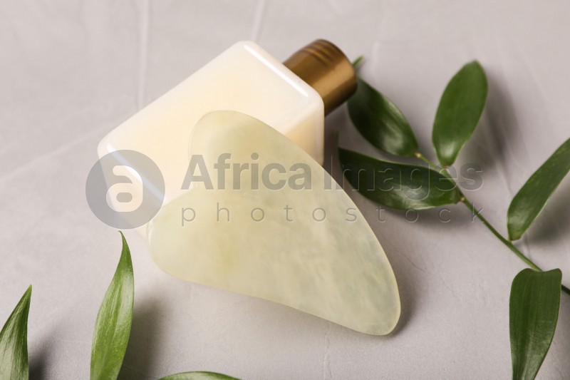 Photo of Jade gua sha tool, cosmetic product and green branches on grey table