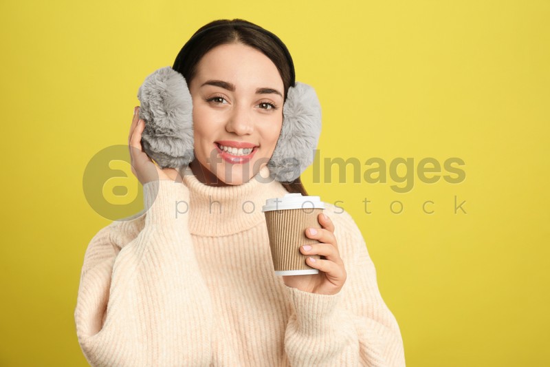 Beautiful young woman in earmuffs with cup of drink on yellow background. Space for text