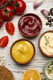 Bowls with mustard, ketchup, mayonnaise and ingredients on wooden table, flat lay