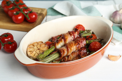 Oven baked asparagus wrapped with bacon in ceramic dish on white wooden table