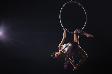 Young woman performing acrobatic element on aerial ring against dark background. Space for text