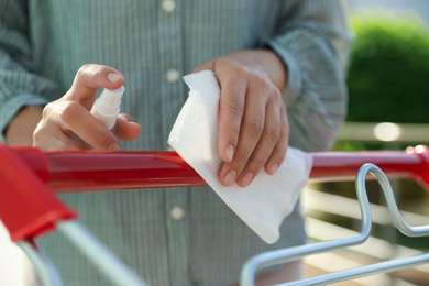 Woman cleaning handle of shopping cart with wet wipe and antibacterial spray on blurred background, closeup