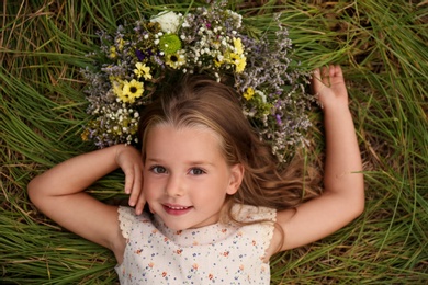 Cute little girl wearing wreath made of beautiful flowers on green grass, top view