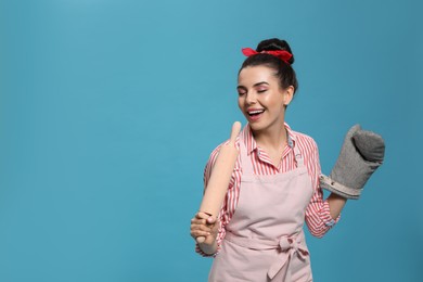 Young housewife in oven glove holding roller pin on light blue background, space for text
