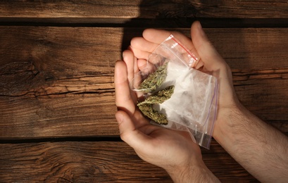 Man holding plastic bags with cocaine and hemp buds on wooden background, top view. Space for text