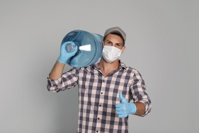 Courier in medical mask with bottle for water cooler showing thumb up on light grey background. Delivery during coronavirus quarantine