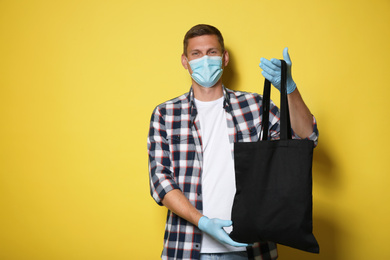 Male volunteer in protective mask and gloves with black bag on yellow background. Aid during coronavirus quarantine