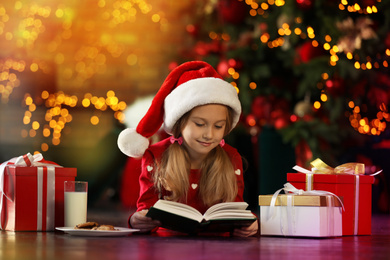 Little girl in Santa Claus cap reading book near Christmas tree indoors