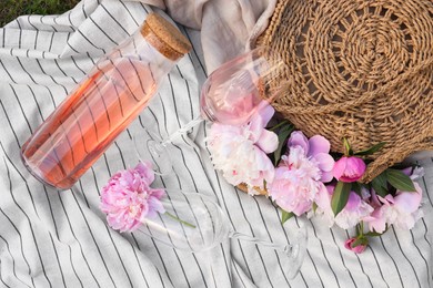 Bottle and glasses of rose wine near straw bag with beautiful peonies on light blanket, flat lay