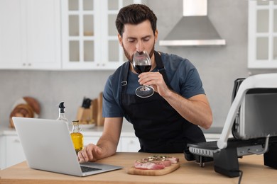 Man with glass of wine making dinner while watching online cooking course via laptop in kitchen