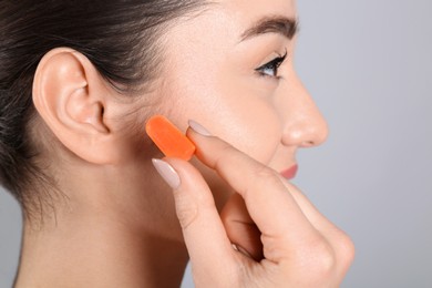 Young woman inserting foam ear plug on grey background, closeup