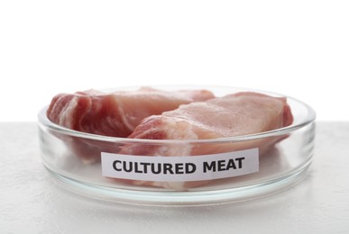 Photo of Petri dish with pieces of raw cultured meat and label on table against white background, closeup