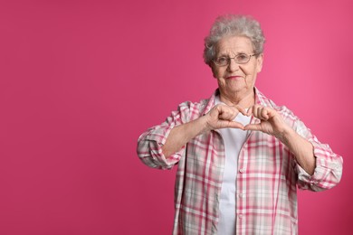 Elderly woman making heart with her hands on pink background, space for text