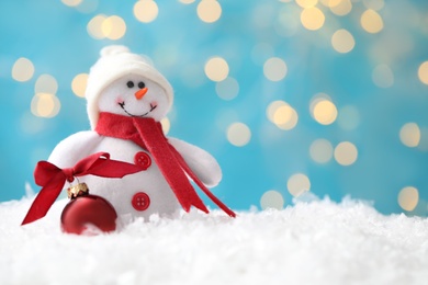 Snowman toy and Christmas ball on snow against blurred festive lights. Space for text