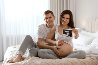 Young pregnant woman and her husband with ultrasound picture of baby in bedroom