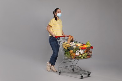 Woman with protective mask and shopping cart full of groceries on light grey background