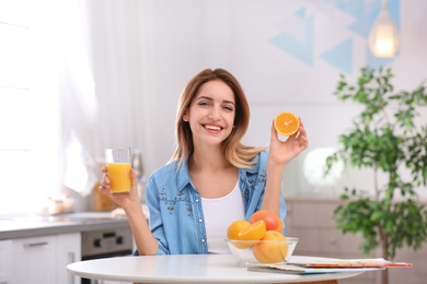 Happy young woman with glass of juice and orange at table in kitchen. Healthy diet