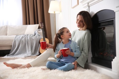 Happy woman and her daughter with cups of hot drink resting near fireplace at home