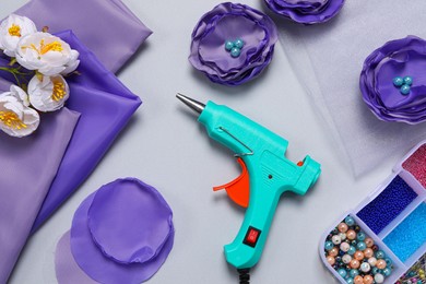 Hot glue gun, textile flowers and handicraft materials on grey background, flat lay