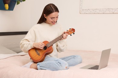 Happy young woman learning to play ukulele with online music course via laptop at home. Time for hobby