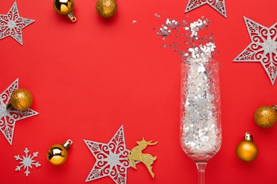Flat lay composition with shiny confetti spilled out of champagne glass near Christmas decorations on red background, space for text