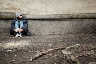 Poor homeless man sitting near wall on street. Space for text