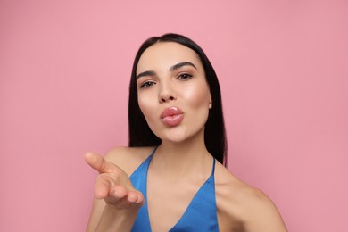 Photo of Beautiful young woman taking selfie while blowing kiss on pink background