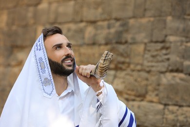 Jewish man blowing shofar on Rosh Hashanah outdoors. Wearing tallit with words Blessed Are You, Lord Our God, King Of The Universe, Who Has Sanctified Us With His Commandments, And Commanded Us To Enwrap Ourselves In Tzitziton in Hebrew