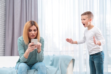 Mother using mobile phone while her son screaming and asking for attention at home. Lonely despaired child