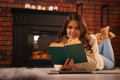 Photo of Young woman reading book on floor near fireplace at home