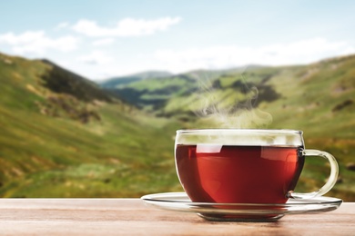Glass cup of fresh hot tea on wooden table against blurred mountain landscape. Space for text