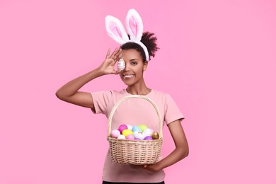 Photo of Happy African American woman in bunny ears headband covering eye with Easter egg on pink background