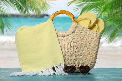 Beach bag with towel, flip flops and heart shaped sunglasses on light blue wooden surface near seashore