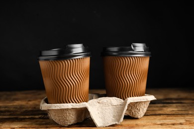 Takeaway paper coffee cups in cardboard holder on wooden table against black background