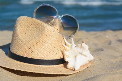 Stylish straw hat, sunglasses and sea shell on sandy beach, closeup