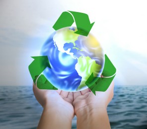 Woman holding virtual image of Earth and recycling symbol near ocean, closeup view