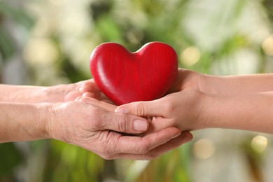 Young and elderly women holding red heart in hands on blurred green background, closeup